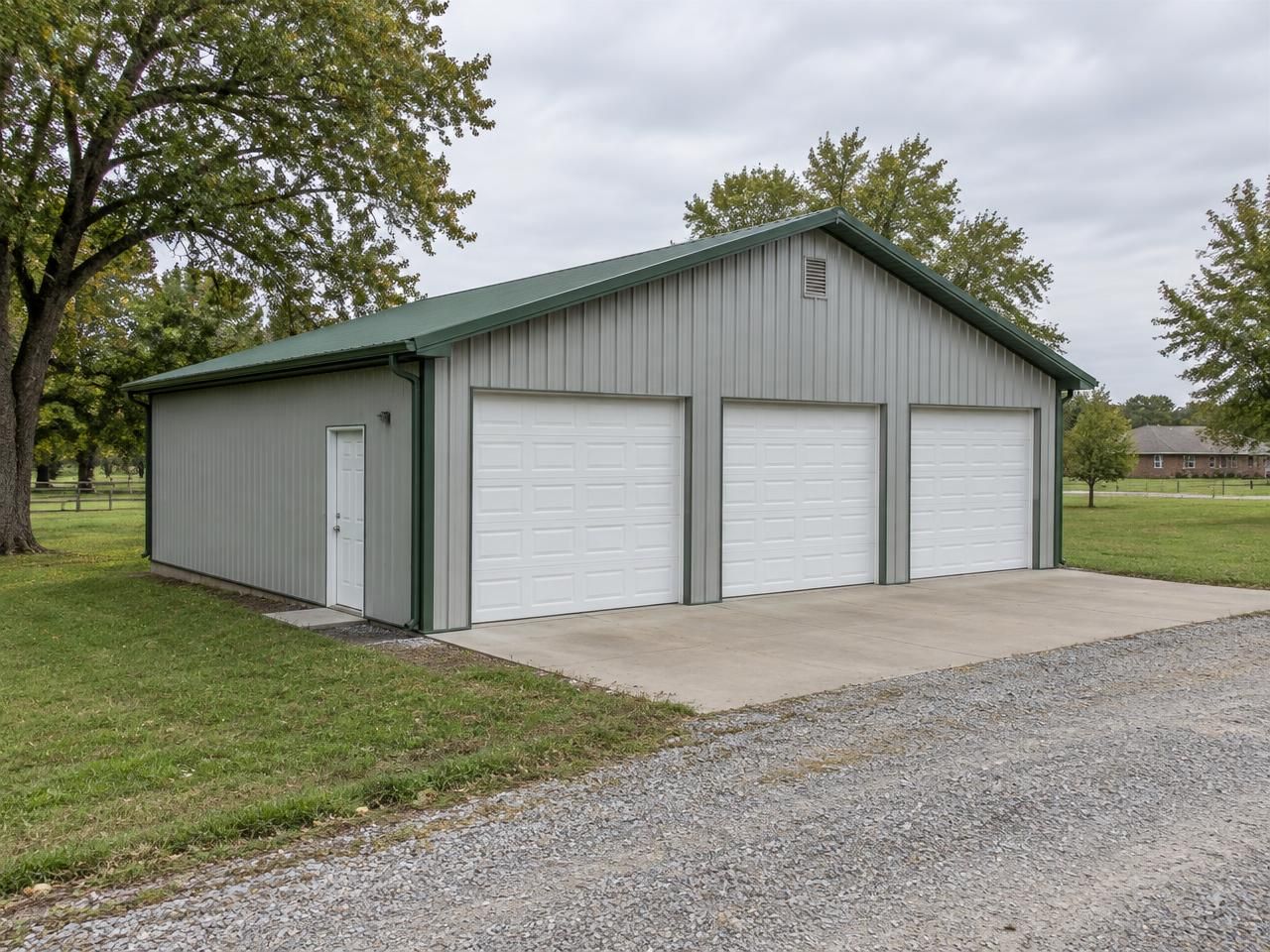 AI-generated illustration: Detached light-gray metal garage with three overhead doors and green roof trim on an Oklahoma-style rural property. Not a completed Outlaw Building Co. project.