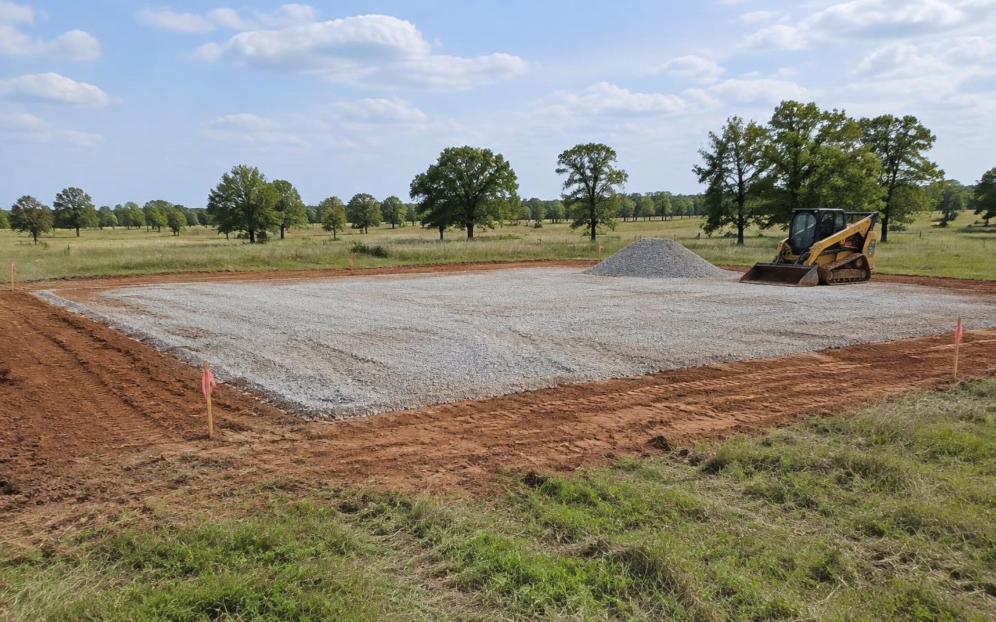 Illustration of a graded gravel building pad with survey stakes and a skid steer on an Oklahoma property